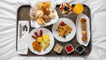 Un plateau de petit-déjeuner contenant divers aliments, notamment des gaufres, des viennoiseries et une salade de fruits, le tout disposé sur un lit.