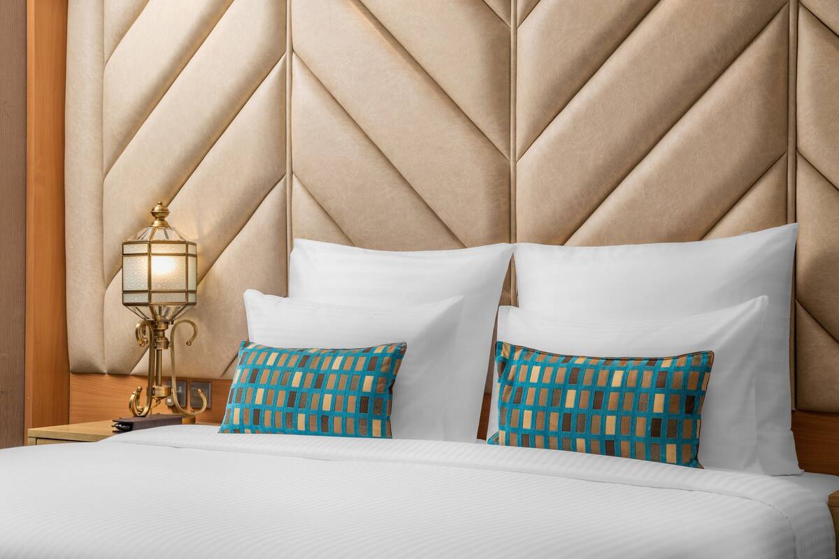 Un lit avec du linge de lit blanc, deux coussins aux motifs bleus et beige, une lampe dorée et une tête de lit en bois ornée d’un motif géométrique.