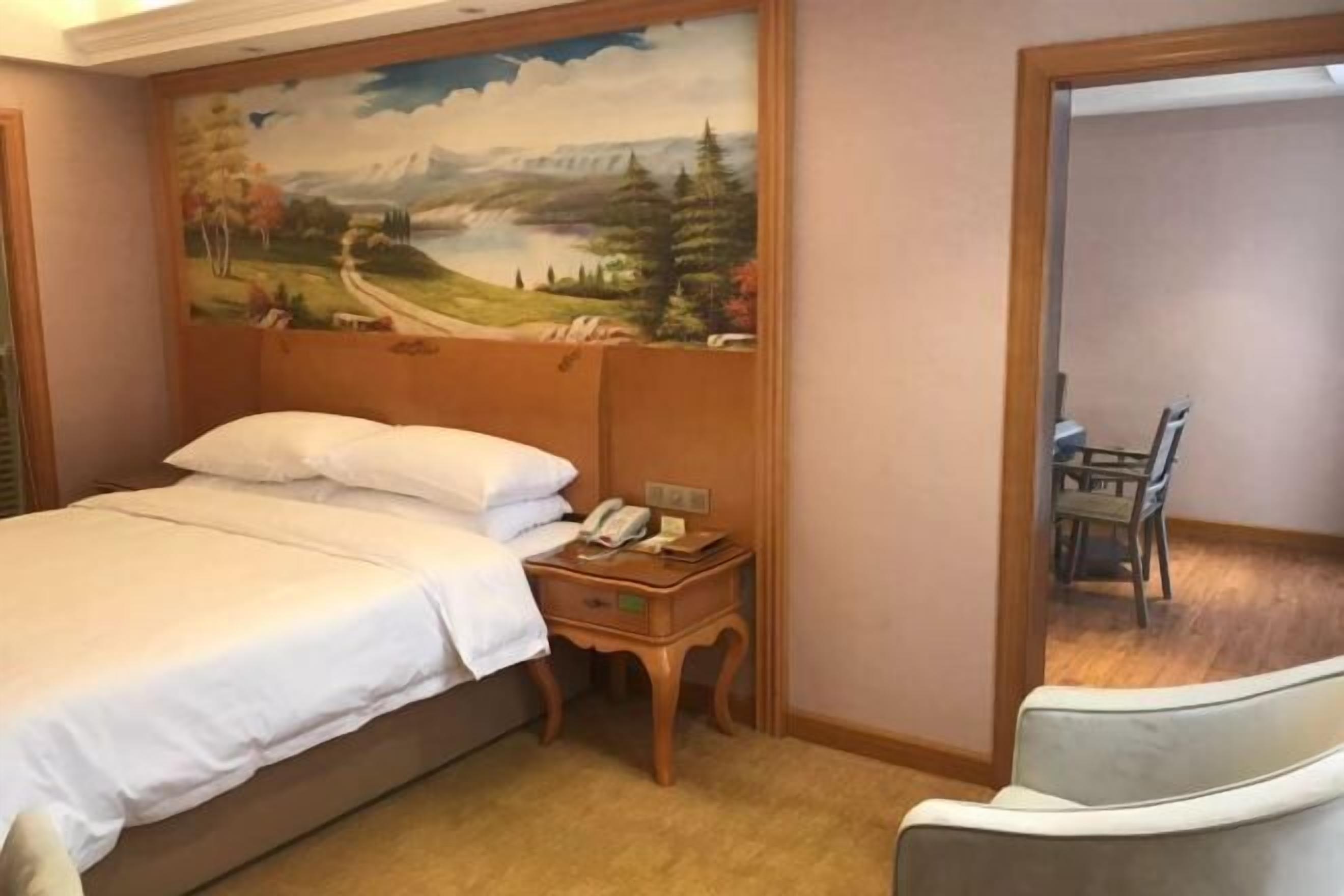 A hotel room with a bed, a nightstand with a telephone, a painting of a landscape on the wall, and a view of a dining area through an open door.