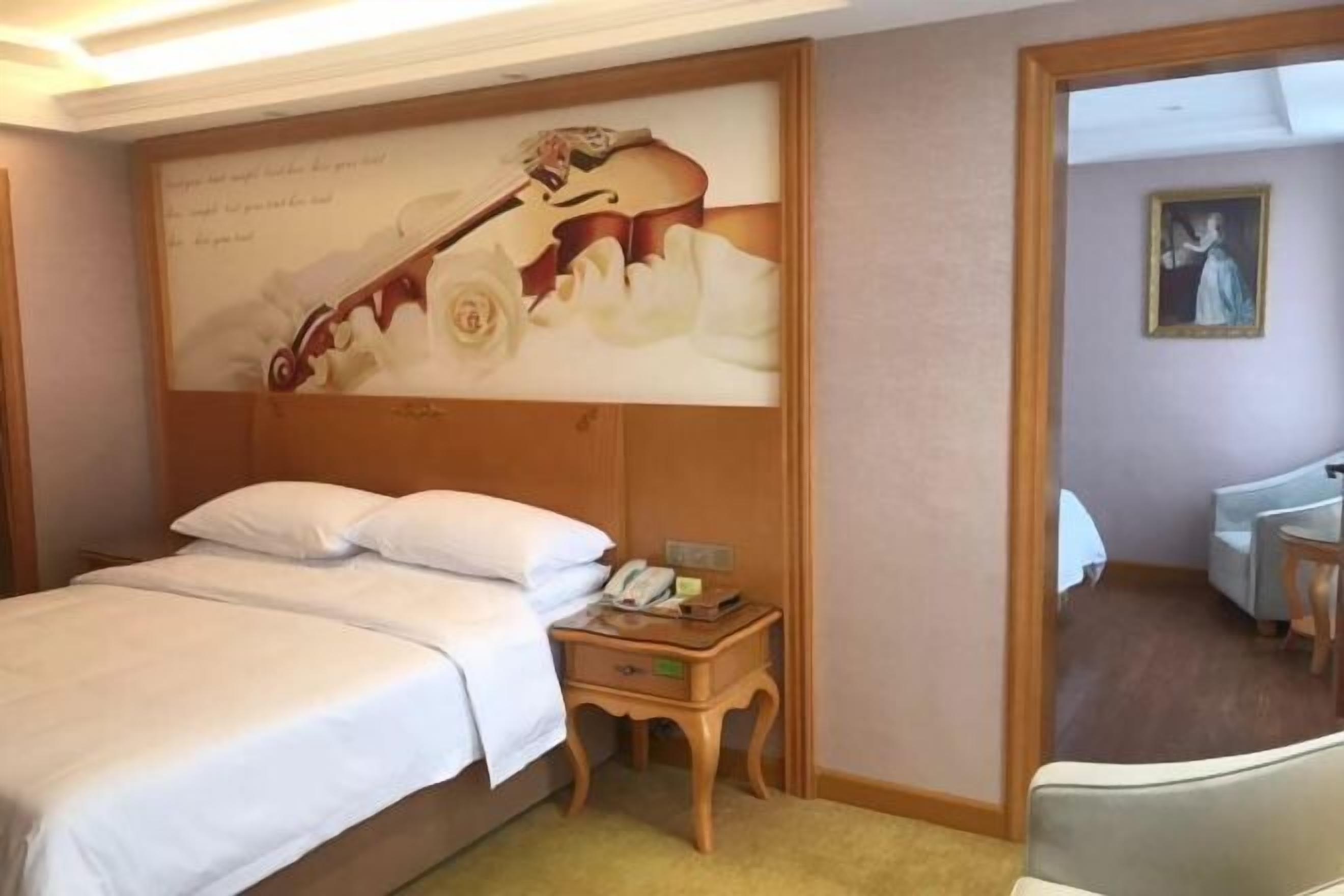 A hotel room with a bed, a nightstand, a painting, and a door leading to another room.