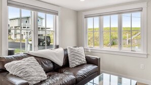 A spacious living room with a brown leather couch, large windows, and a view of a residential area. - The Nest | High-End Kitchen | Steps to Beach | A/C (Newbury)