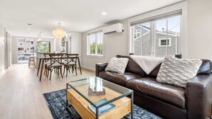 A modern living room with a glass coffee table, a brown leather sofa, and a dining area with wooden chairs. - The Nest | High-End Kitchen | Steps to Beach | A/C (Newbury)