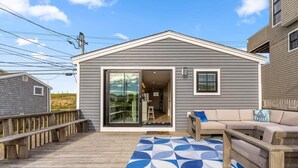 A covered deck with a blue and white patterned rug, a wooden bench, and a sofa with cushions. - The Nest | High-End Kitchen | Steps to Beach | A/C (Newbury)