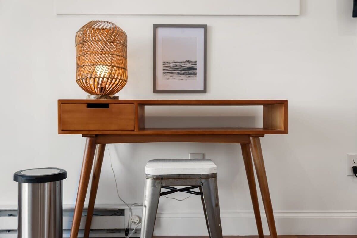A wooden desk with a wicker lamp, a framed picture, and a metal stool.