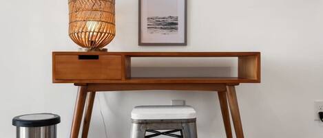 A wooden desk with a wicker lamp, a framed picture, and a metal stool.