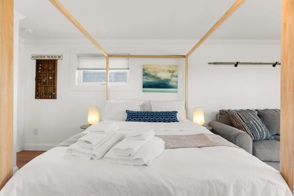 Una cama bien hecha con sábanas blancas y toallas dobladas, un sofá gris con cojines estampados, un cabecero de madera, un estante montado en la pared con objetos decorativos y un cuadro sobre la cama.