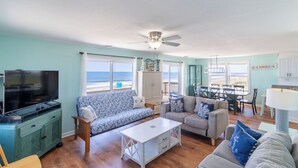 A spacious living room with a large TV, two sofas, a dining area, and a view of the beach. - Sweeping Ocean Views, Spectacular Sunrises, One-level living, KD3311 (Kill Devil Hills)