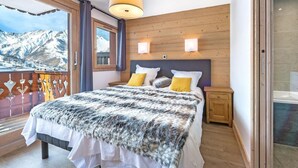 Quarto com uma cama grande, cabeceira de madeira, duas mesas de cabeceira e vista para montanhas cobertas de neve através de uma varanda.