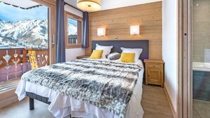 Ein Schlafzimmer mit einem großen Bett, einem Holz-Kopfteil, zwei Nachttischen und Blick auf schneebedeckte Berge vom Balkon aus.