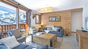 Ein gemütliches Wohnzimmer mit einem großen Fenster, Holzwänden und einem Balkon mit Blick auf schneebedeckte Berge.