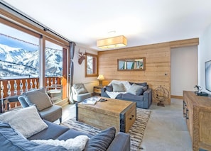 Ein gemütliches Wohnzimmer mit einem großen Fenster, Holzwänden und einem Balkon mit Blick auf schneebedeckte Berge.
