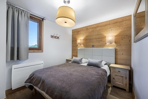 A bedroom with a bed, bedside tables, a window with curtains, a wooden wall panel, and a ceiling light fixture.