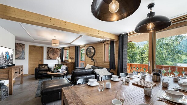Un salon chaleureux avec un plafond en bois, un canapé en cuir noir, une table à manger en bois mise avec des assiettes, des tasses et des couverts, et une grande fenêtre donnant sur un paysage verdoyant.