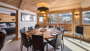 Un interior de madera con una mesa de comedor dispuesta para seis personas, una cama con cabecera y un ventanal con vistas a un paisaje nevado.
