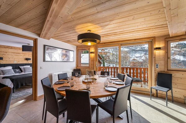 Un interior de madera con una mesa de comedor dispuesta para seis personas, una cama con cabecera y un ventanal con vistas a un paisaje nevado.