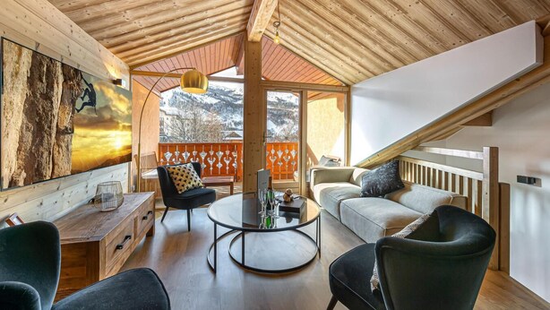 Un salón espacioso con techo de madera, un ventanal grande con vistas a una montaña nevada, un sofá, una mesa de centro y un televisor montado en la pared.