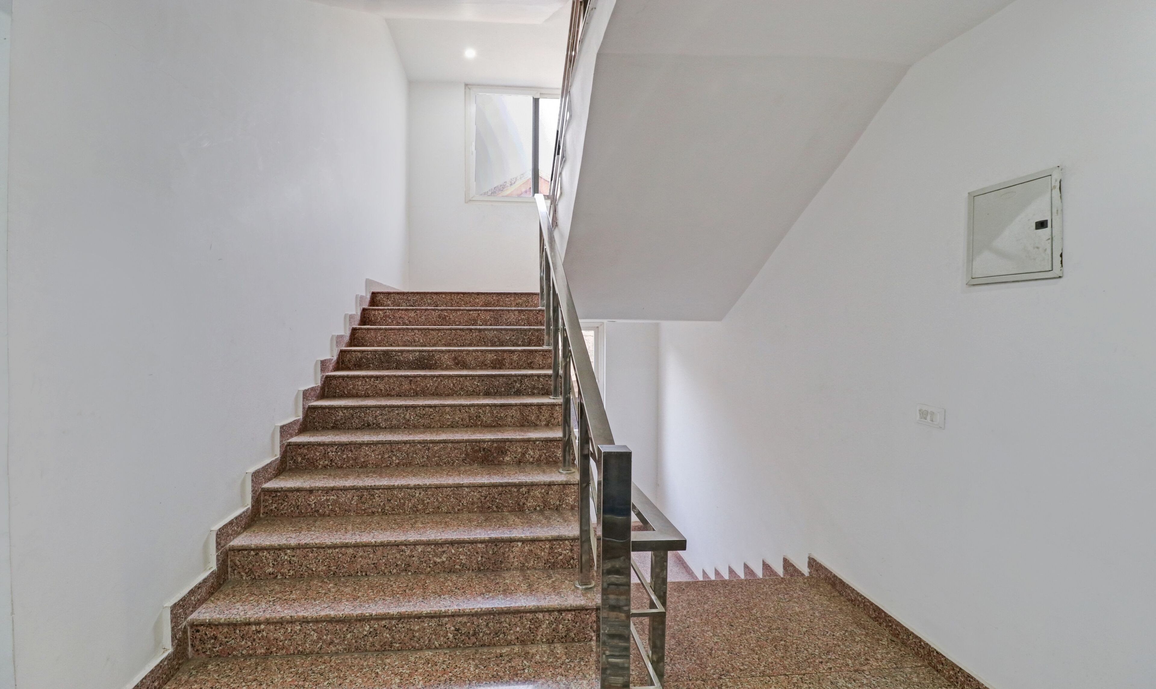 A staircase with granite steps and metal railings, leading to a landing with a white wall and a small window.
