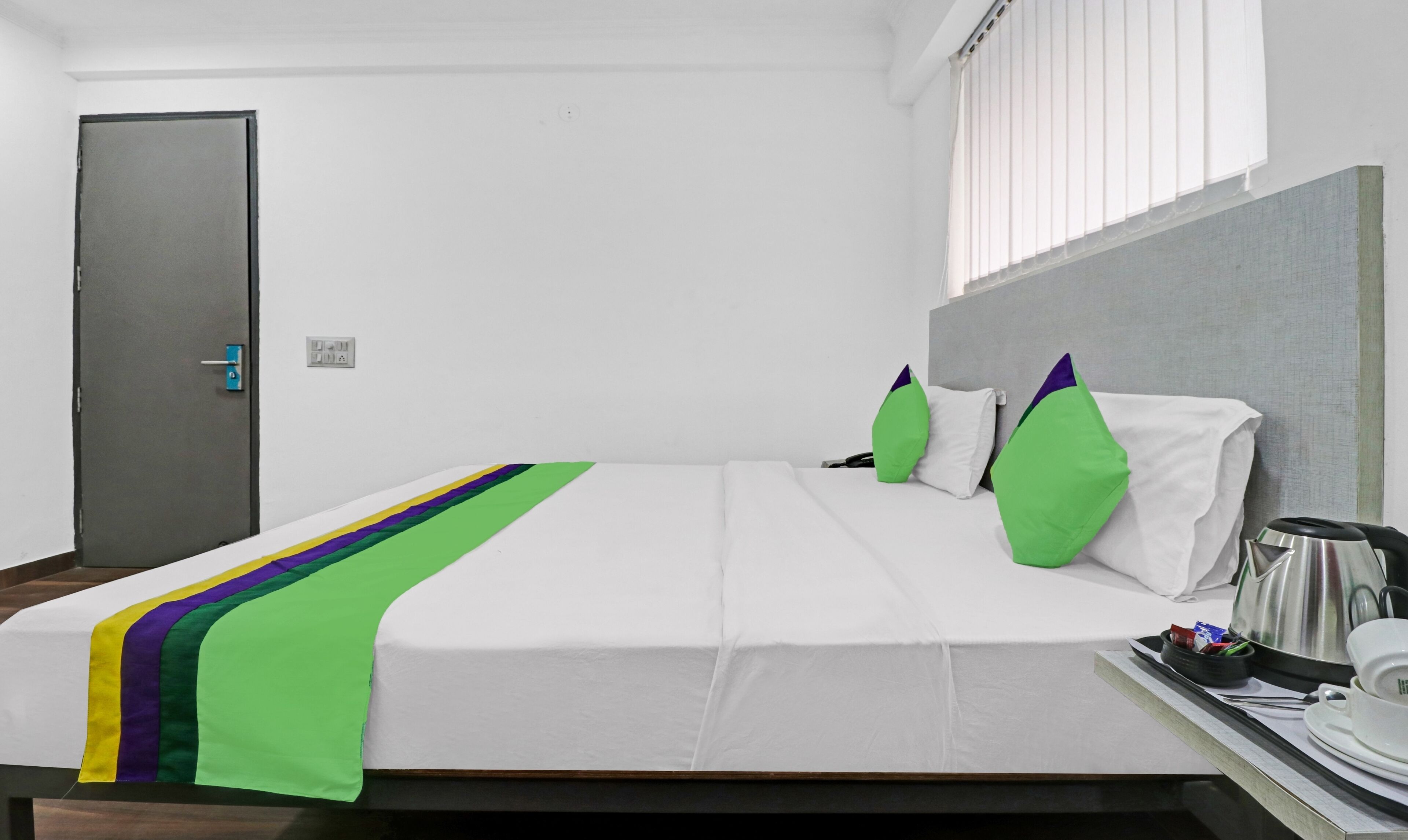 Une chambre d’hôtel avec un lit, une fenêtre avec des stores, une porte et un plateau avec une bouilloire et des tasses.