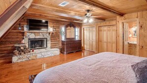 Une chambre située dans une cabane en rondins, avec une cheminée en pierre, des poutres apparentes en bois au plafond, un ventilateur de plafond et un grand lit.