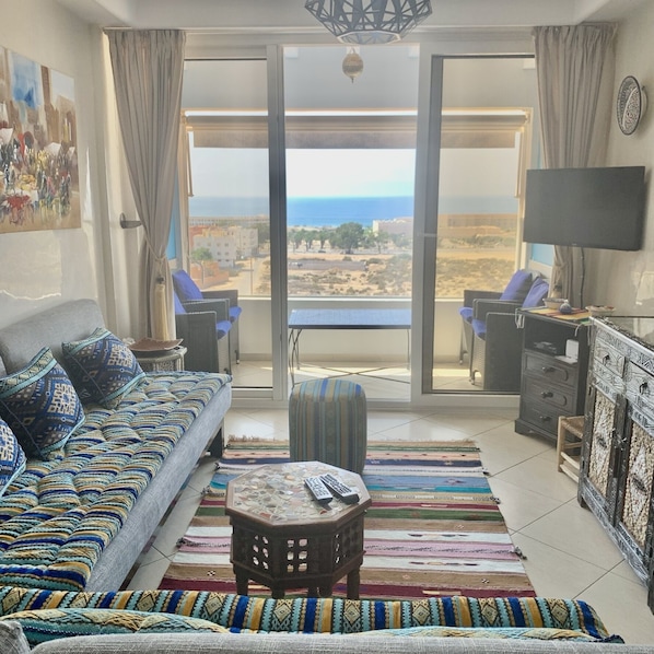 A living room with a sofa, a small table, a TV, and a view of the sea through large glass doors.