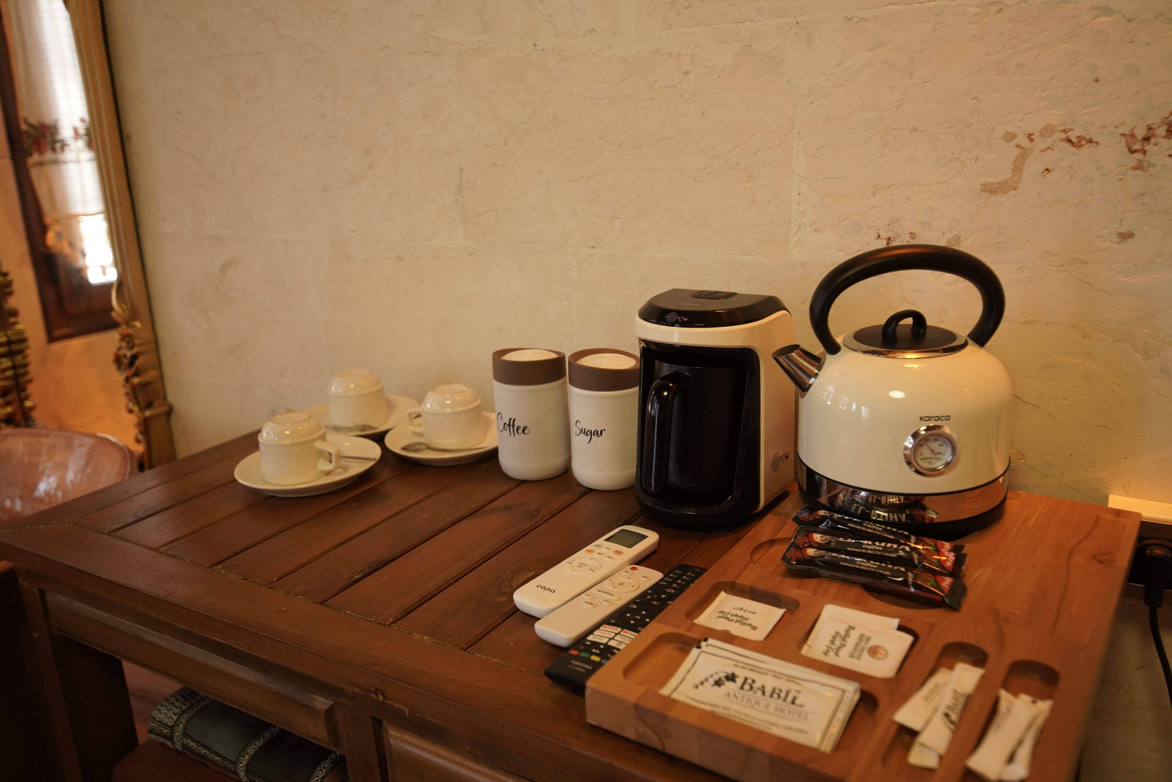 A wooden table with a kettle, coffee maker, cups, sugar and coffee containers, remote controls, and a box of baby wipes.