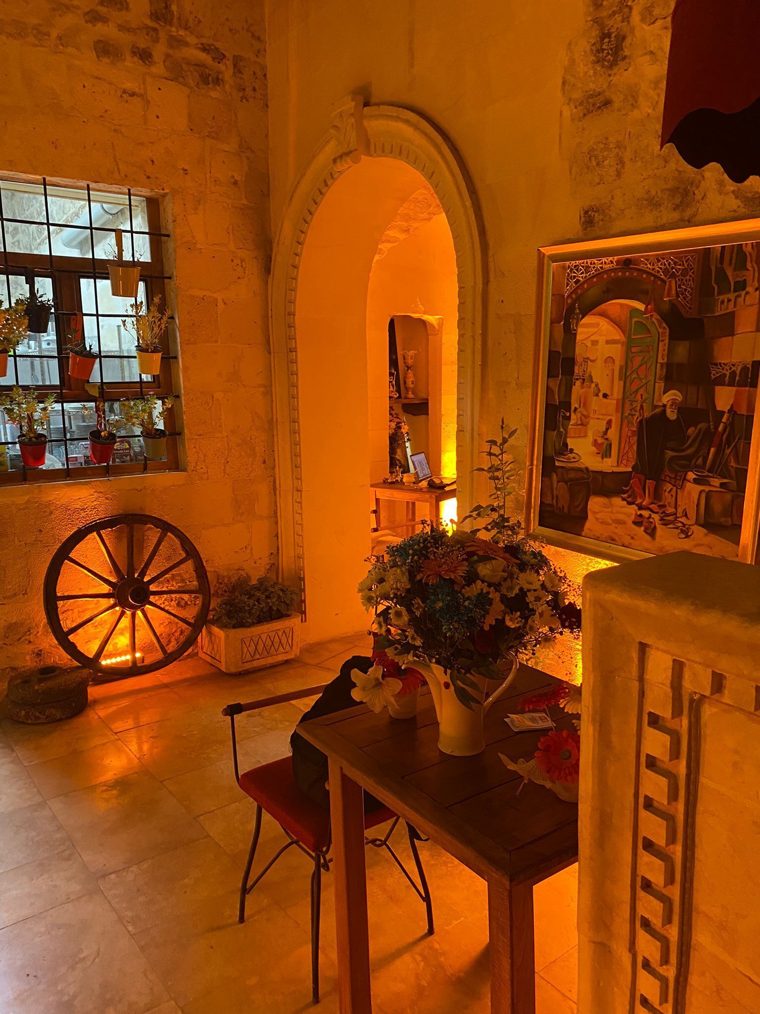A room with a large wooden wheel, a table with a vase of flowers, and a window with potted plants.