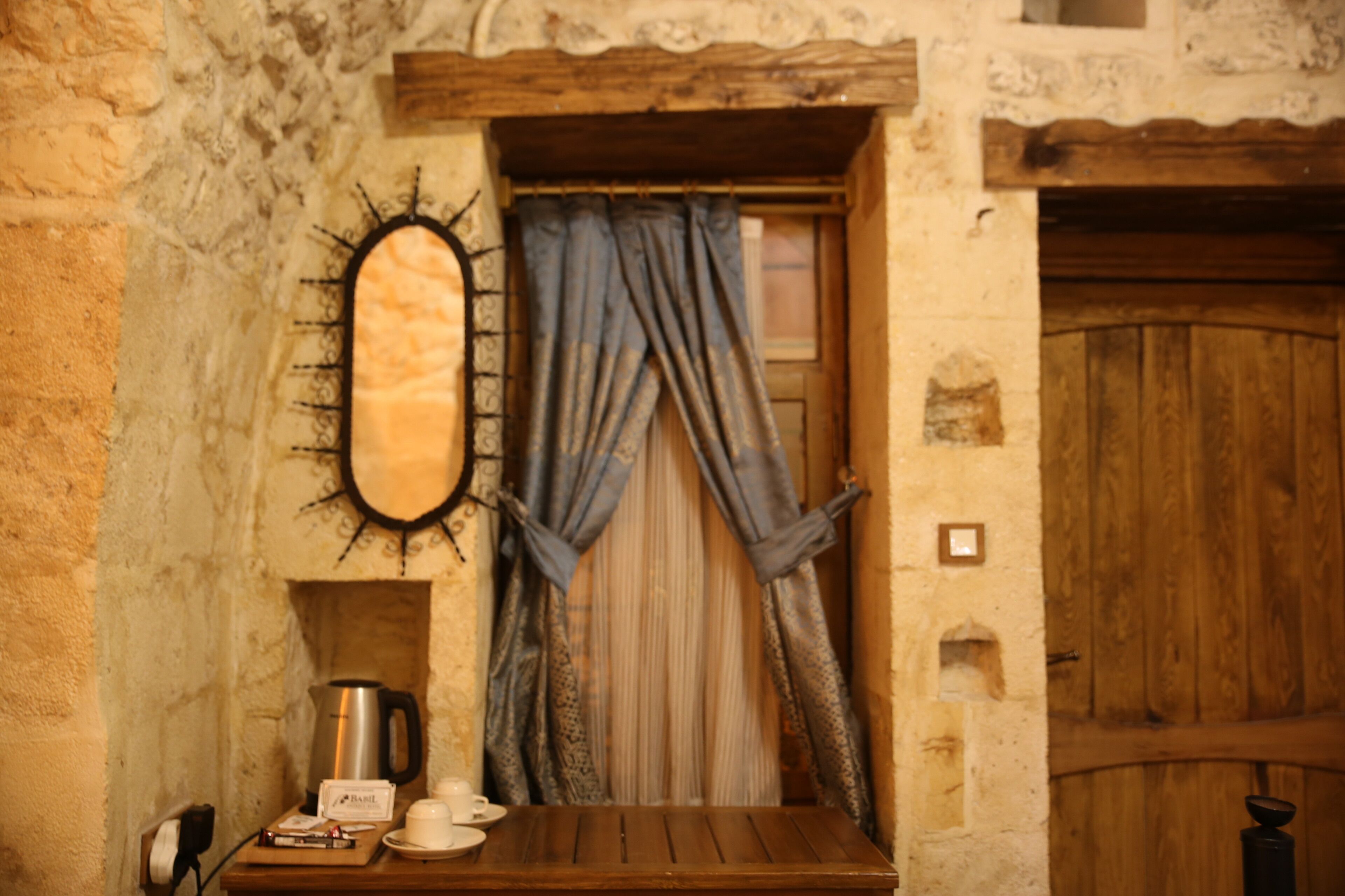 Una stanza rustica con un tavolo di legno, un bollitore, una tazza e un cartello con su scritto 'Barbe'. Si intravede una finestra con tende, e sullo sfondo una porta in legno.