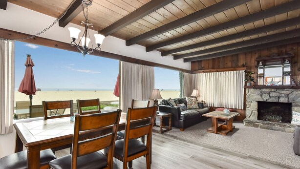A spacious living area with a view of the beach, wooden ceiling beams, a stone fireplace, and a dining area with chairs and a table. - West Newport Oceanfront Home & Patio on 65th St. (Newport Beach)