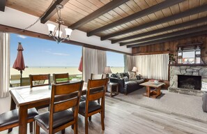Un'ampia zona giorno con vista sulla spiaggia, travi in legno al soffitto, un camino in pietra e un'area pranzo con sedie e tavolo.