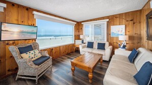 Una habitación con interiores de madera, un ventanal grande con vistas a la playa, una silla de mimbre, un sofá blanco y una mesa de centro de madera.
