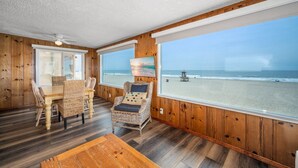 Una habitación con interior de madera, una mesa de comedor con sillas, un sillón de mimbre con cojines y ventanas amplias que dan a una playa.
