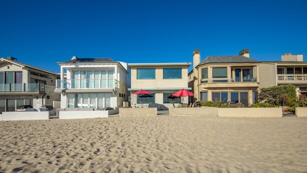 Una propiedad frente a la playa con una arena dorada, un área de piscina con sombrillas y casas modernas con amplios ventanales.