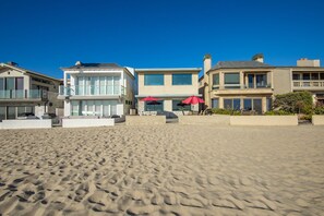 A beachfront property with a sandy beach, a pool area with umbrellas, and modern houses with large windows.