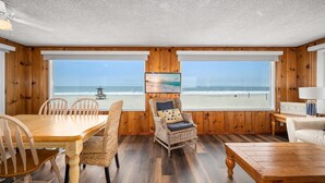 Una habitación con interior de madera, ventanas amplias con vistas a la playa, una mesa de comedor, sillas, un sillón de mimbre y un pequeño sofá.
