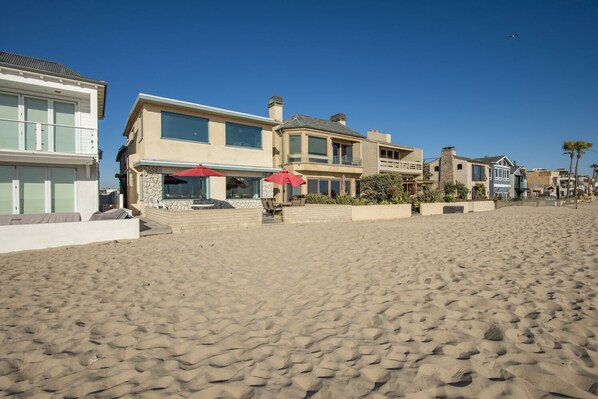 A beachfront property with a large house featuring multiple windows, a patio with red umbrellas, and a sandy beach in the foreground.