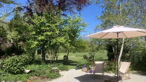 Un patio con suelo de grava, rodeado de abundante vegetación y árboles. Se observan dos sillas de madera bajo un gran paraguas.
