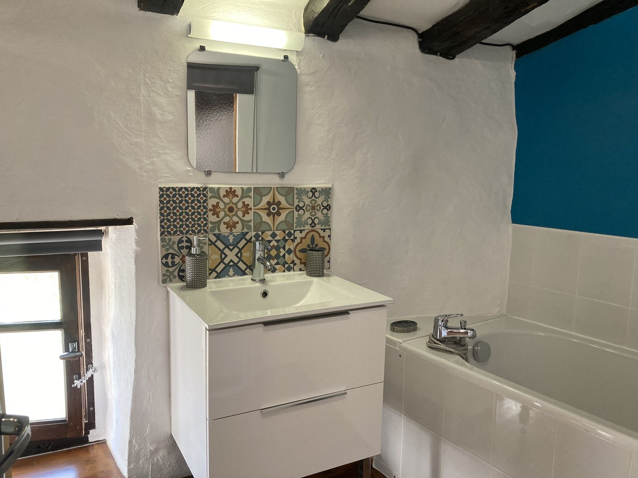 Een badkamer met een wit wastafelmeubel, een spiegel, een betegelde achterwand met patroon, een witte kast en een bad.
