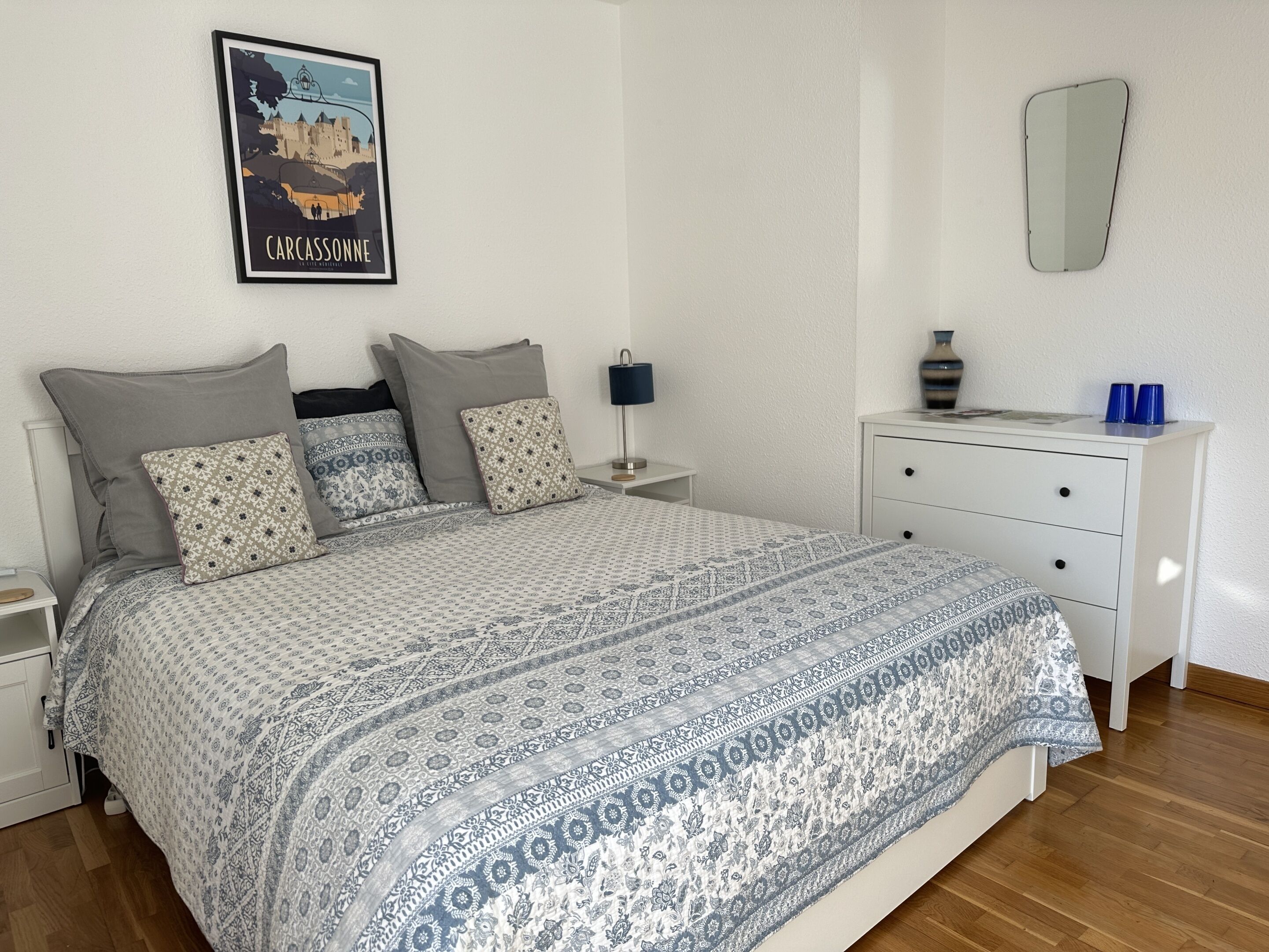 A bedroom with a patterned bedspread, a nightstand with a lamp, a dresser, and a poster on the wall.