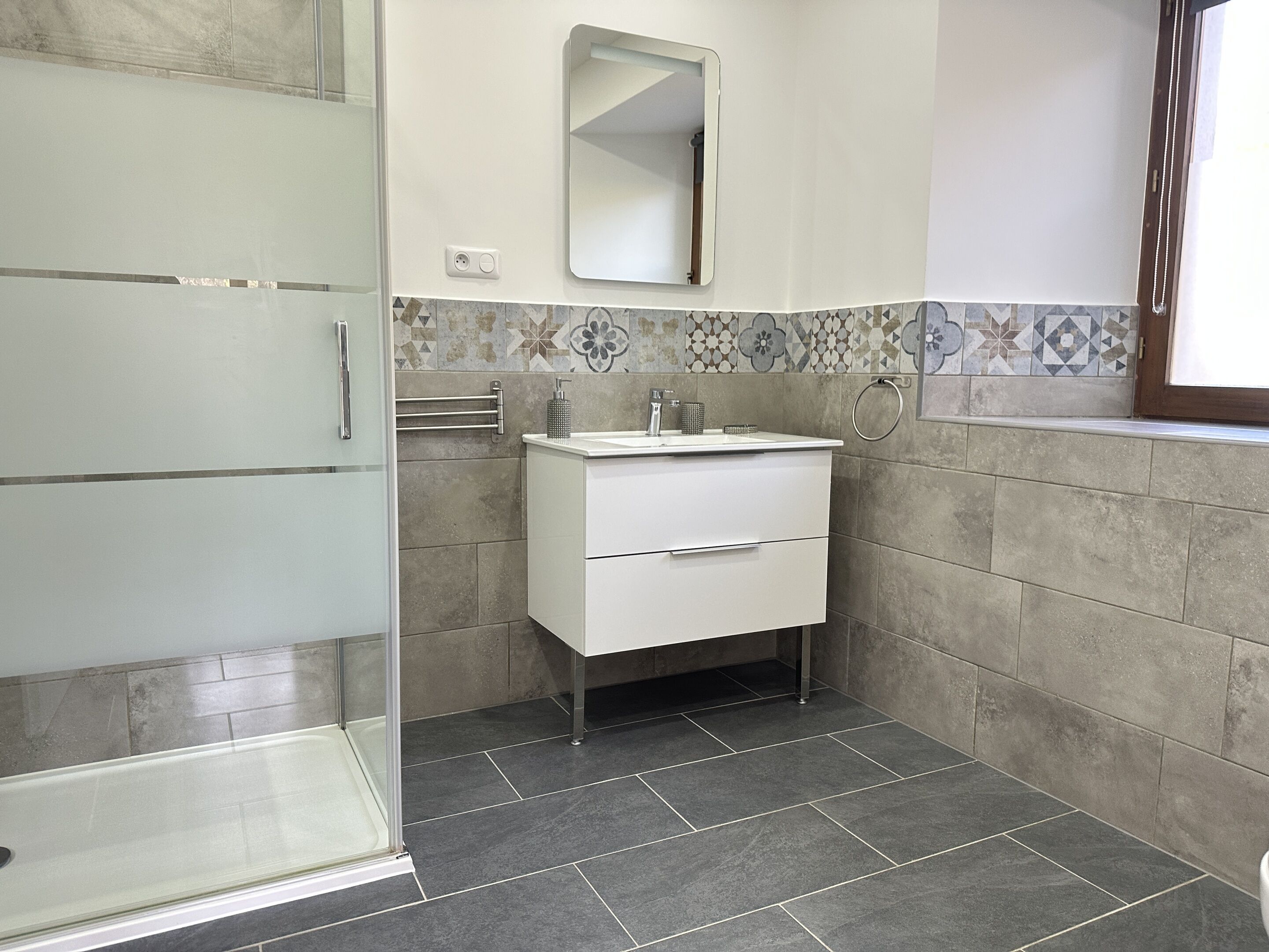 Een moderne badkamer met een douchecabine van glas, een witte wastafel met een spiegel erboven en behangtegels op de muren.