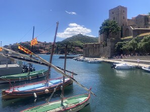 Un port où sont amarrés des bateaux, une structure ressemblant à un château et une colline avec des bâtiments en arrière-plan.