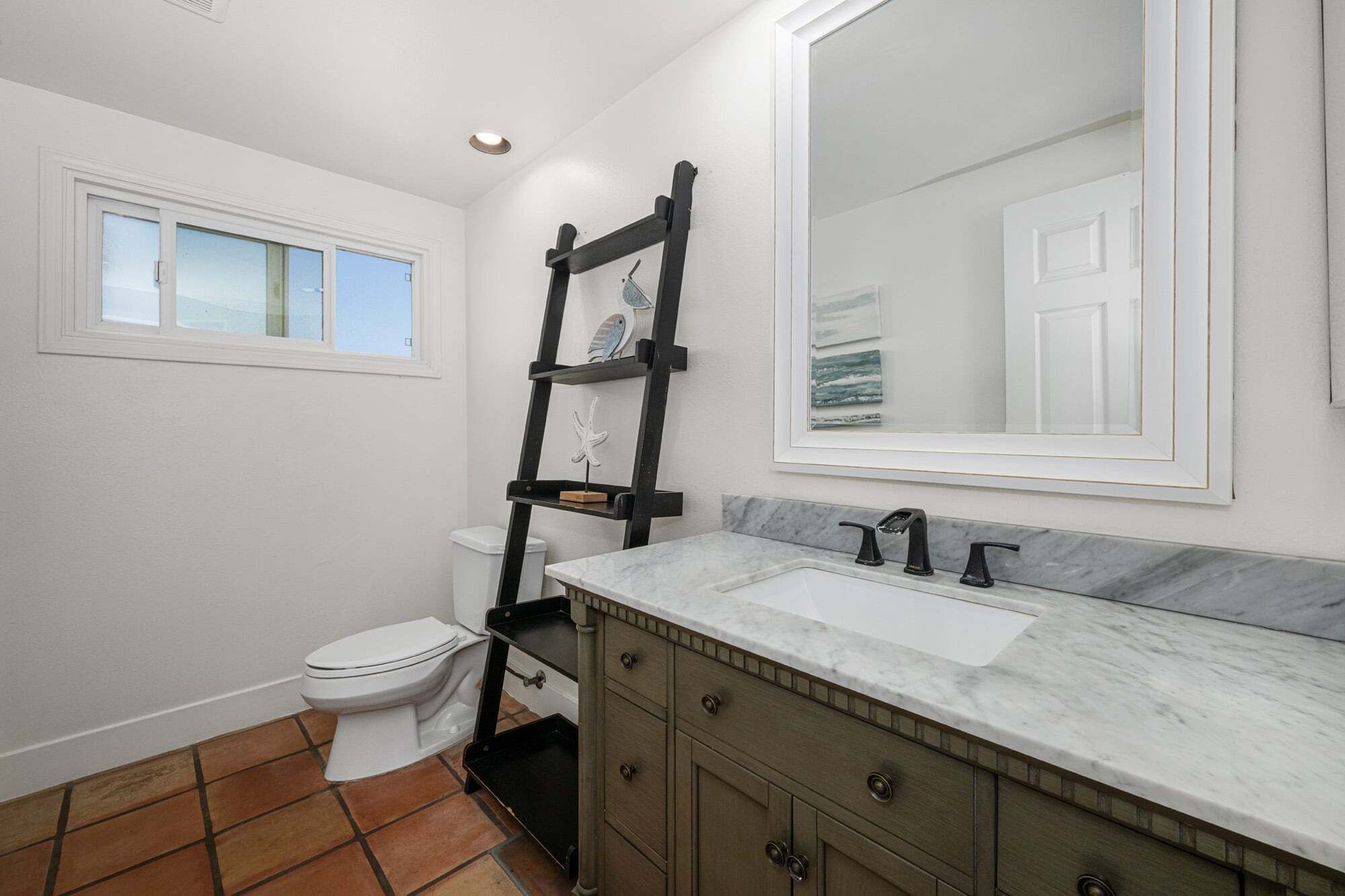 Un baño con inodoro, lavamanos con encimera de mármol, tocador de madera, espejo y ventana.
