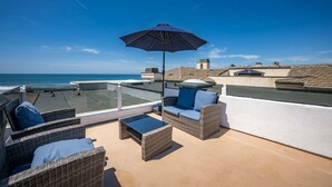 Eine Dachterrasse mit Korbmöbeln, einer Glasbrüstung und Blick auf das Meer.