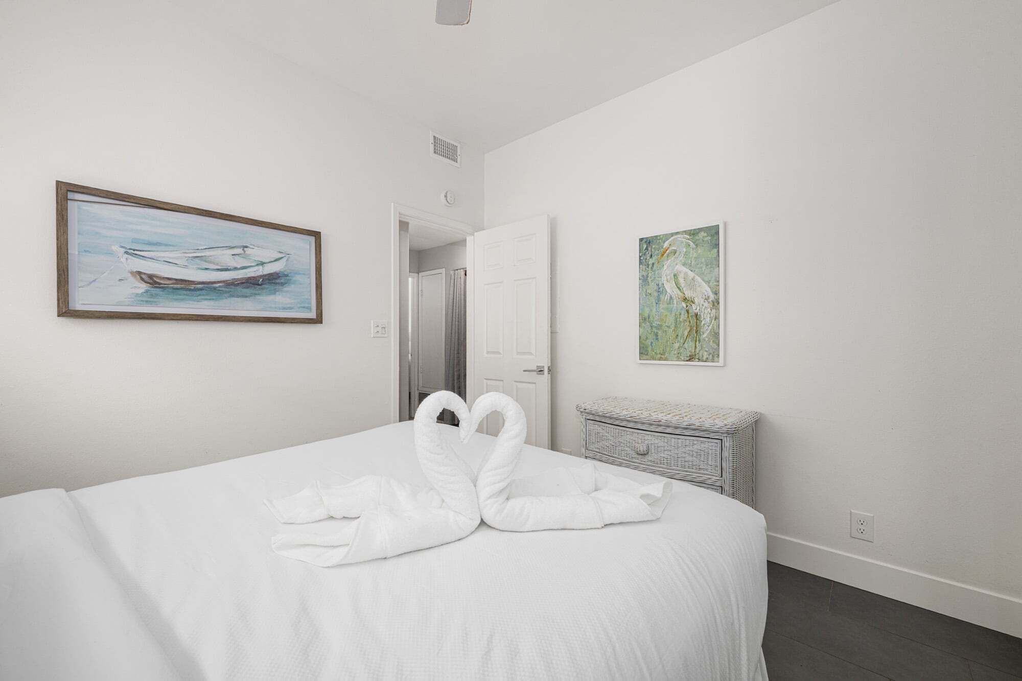 A bedroom with a bed, a painting of a boat, a painting of a bird, and a wicker chest.