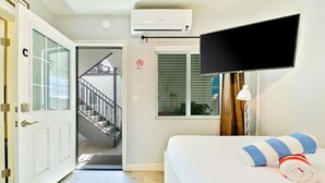 Ein Hotelzimmer mit Bett, Fernseher, Klimaanlage, einem Fenster mit Jalousien und einer Treppe, die in einen Außenbereich führt.
