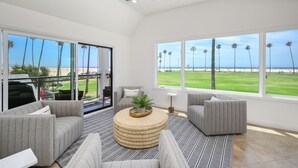 Een ruime woonkamer met een groot raam met uitzicht op een strand, een plafondventilator en een ronde salontafel met een plant.