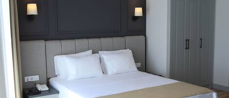 A neatly made bed with white linens and a beige throw blanket, a bedside table with a phone and charger, and a wardrobe with two doors.
