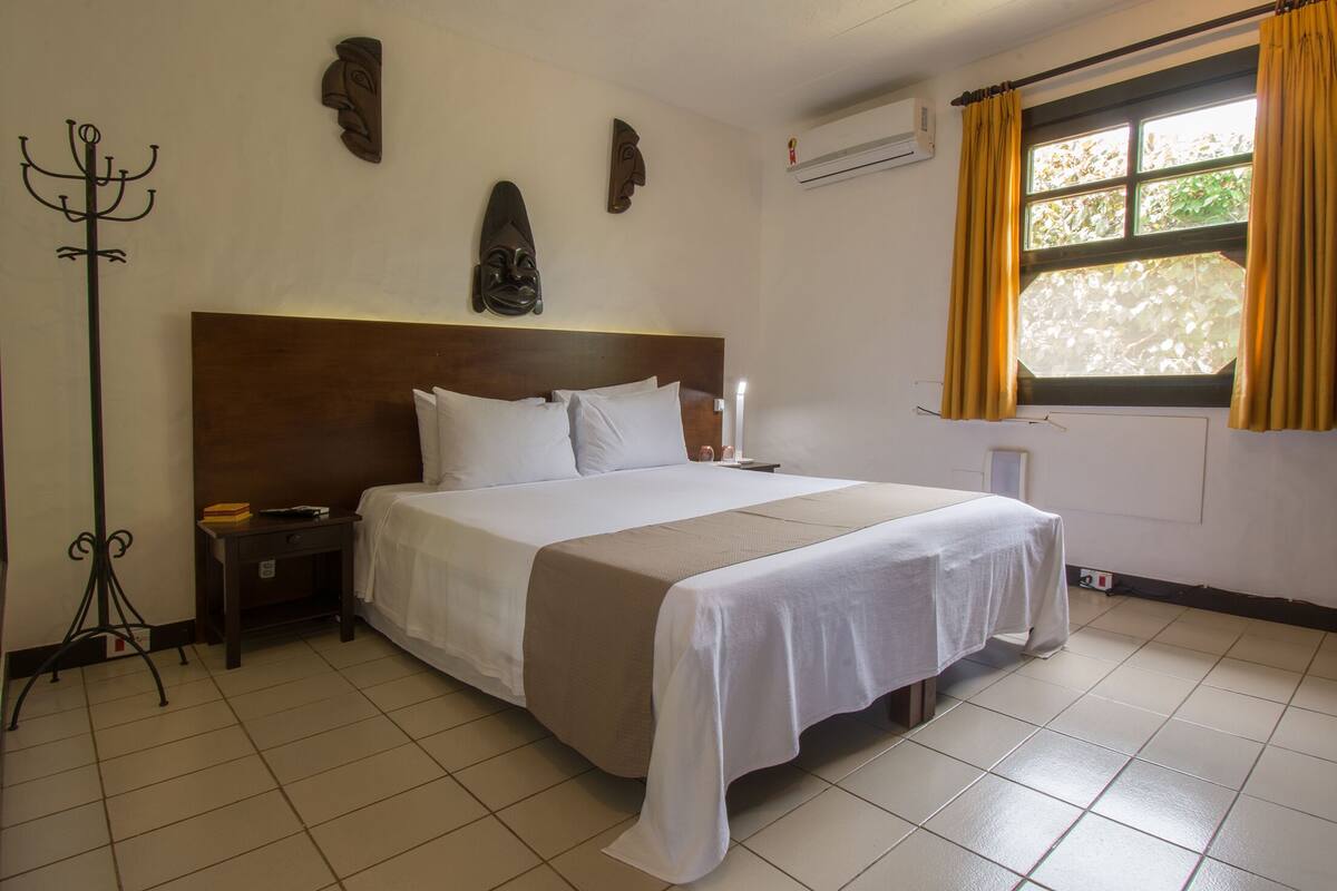Une chambre d’hôtel avec un grand lit, une table de chevet, un porte-manteau et une fenêtre avec des rideaux.