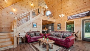 A wooden cabin interior with a red leather sofa, a wooden coffee table, a staircase with a railing, and a kitchen area.
