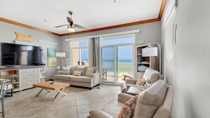 Een ruime woonkamer met een grote flatscreen-tv, een plafondventilator en uitzicht op de oceaan door grote glazen schuifdeuren.