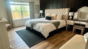 Une chambre à coucher comprenant un lit, des tables de chevet, une commode, une chaise, un bureau, un miroir, un ventilateur de plafond et une fenêtre avec des rideaux.